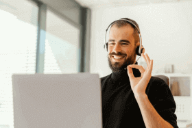 Un hombre con barba haciendo una seña de excelente con su mano, frente a su laptop