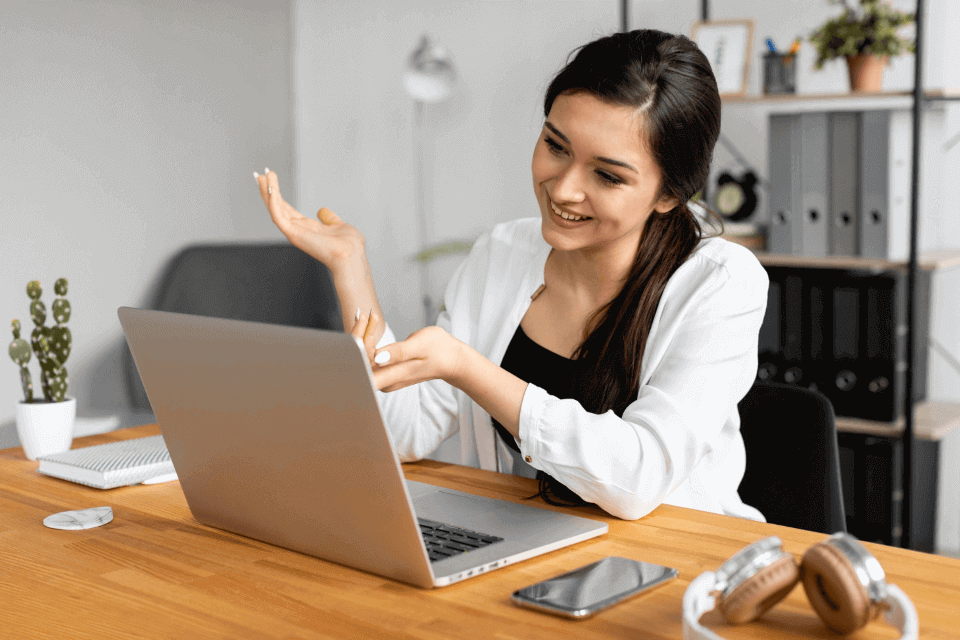 Una mujer sentada en su escritorio, en una oficina, frente a su laptop, sonriendo y viendo sus cursos de rrhh online.  