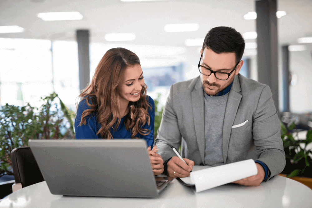 Una mujer y un hombre sentados en un escritorio, frente a una laptop, revisando un documento sobre el feedback 360 grados.